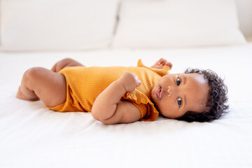 Obraz premium Funny little African American baby girl in orange bodysuit on the bed at home on the back, funny six-month-old black newborn baby close-up
