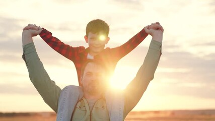 Happy father carries son on shoulders at back sunset light. Dad and boy take leisurely walk in midst of nature spending time together on sunny day. Father with young boy shares happiness