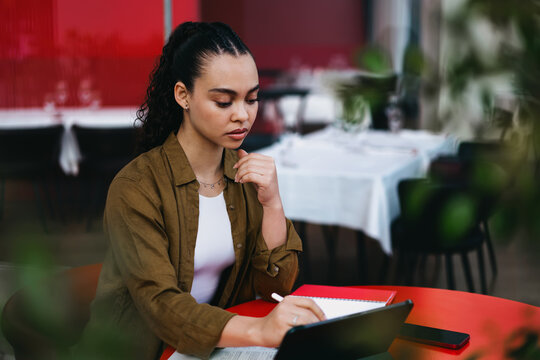 Thoughtful Hispanic woman working remotely in stylish café, use tablet, writing in notebook. Independent professional managing business projects,studying, planning strategies. Digital entrepreneurship - Powered by Adobe