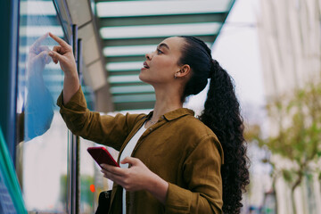 Young Latina woman interacts with touchscreen information board at public transportation stop....
