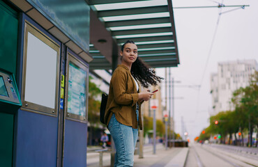 Confident young woman with flowing hair stands at tram stop, holding phone and waiting for public transport. motion in her hair adds dynamism, symbolizing movement, freedom, and urban exploration