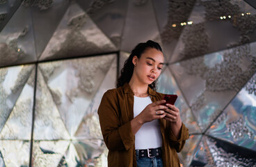 Confident Latina woman browsing her smartphone while standing in urban setting with reflective metallic background. Female embodies digital lifestyle, social media engagement, and smart connectivity
