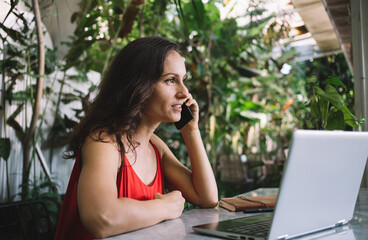 Focused middle-aged woman in red dress speaking on the phone while working on her laptop in a lush,...