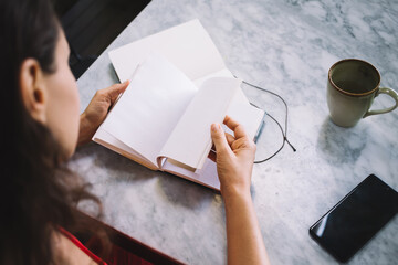 Woman flipping through pages of a book on a marble table with a coffee mug, smartphone, and open...