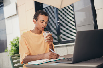Young man enjoying a refreshing drink while studying at an outdoor cafe on a sunny day in the city