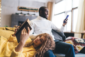 Relaxed couple at home, enjoying leisure time on the couch. Dark-skinned man checks banking app on smartphone while woman rests on his lap, reading a book. Cozy, warm atmosphere peaceful connection