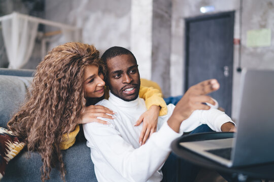 Couple shopping online at home, sitting on the cozy couch with laptop. The dark-skinned man points at the screen while his partner embraces him, both looking engaged and happy in a relaxed setting