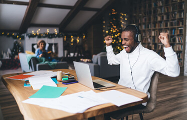 Fototapeta premium Joyful African American man in headphones celebrating success at home, raising arms in excitement while working on laptop. Cozy holiday setting with festive decor and cheerful atmosphere