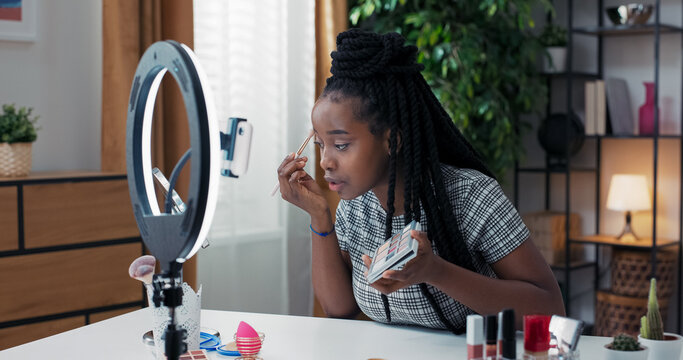 Beauty blogger woman filming daily eye makeup routine tutorial near phone camera on tripod. Influencer girl live streaming cosmetics product review. - Powered by Adobe