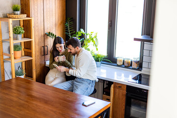 Young couple sitting in their modern kitchen, enjoying a cup of coffee and browsing social media on their smartphone