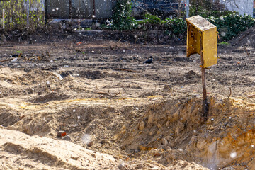 construction site with excavated earth and a lonely yellow gas box on a metal post. falling snow spring.