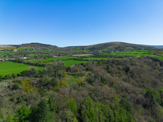 A wide aerial shot captures a vast landscape of rolling green hills, interspersed with fields, trees, and distant houses under a clear blue sky.