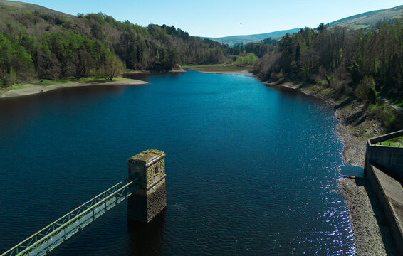 Aerial view of a deep blue reservoir surrounded dense woodland. A stone Tower with a walkway extends into tranquil water.