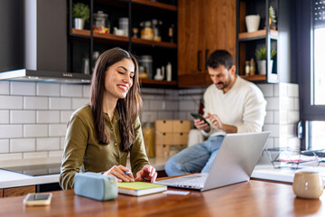 Young woman working or studying from home using laptop in kitchen while man using phone in background