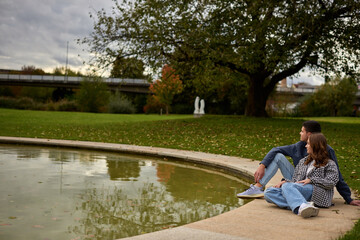 Romantic young couple relaxing by the pond in autumn park, wearing cozy casual outfits, enjoying a peaceful moment together.