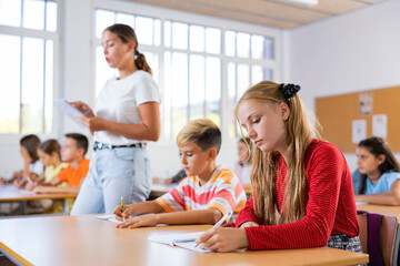Portrait of interested diligent preteen schoolgirl sitting at school desk and writing exercises at lessons with classmates