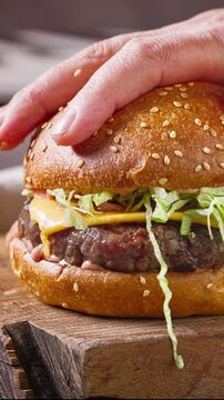 cook put chopped lettuce on grilled burger meat for making delicious cheeseburger on wooden cutting board, food closeup