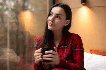 A young woman in red plaid pajamas sits by a window, holding a black mug. She gazes thoughtfully...