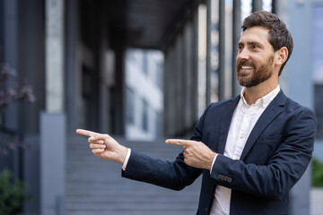 A smiling businessman in a dark suit points both fingers to the right, near an office building,...