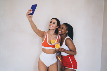 Playful athletic women with fresh orange slices smiling at front mobile camera during together...