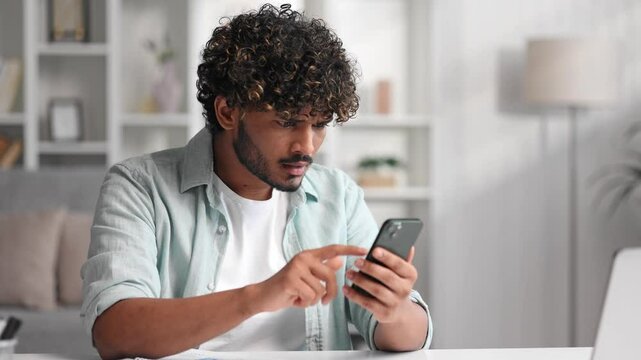 Worried young Indian boy sitting on sofa looking at her phone and does not understand why it is not working. The angry student in glasses does not believe the bad news he saw.