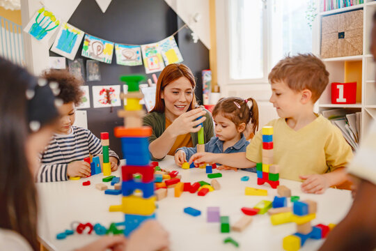Preschool teacher interacts with a diverse group of children building with colorful blocks in a bright classroom, promoting creativity, teamwork, and multicultural early education.