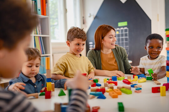 Preschool teacher interacts with a diverse group of children building with colorful blocks in a bright classroom, promoting creativity, teamwork, and multicultural early education.