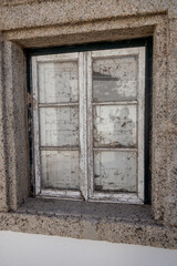 Old wooden window with peeling paint and dirty glass set in a granite stone wall, captured during a daytime urban exploration to document architectural decay in a historical village.