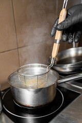 A person in black gloves holds a wire skimmer over a steaming pot of boiling water on a stove. The scene captures the process of preparing food in a professional kitchen setting.