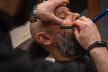 Obraz premium Close-up of a barber trimming a beard with electric clippers in a modern barbershop. Professional men's grooming and beard care service, selective focus