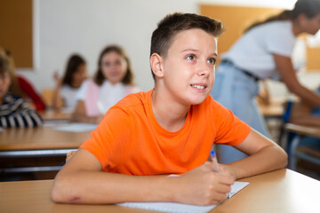 Portrait of concentrated tween boy studying in classroom, listening to schoolteacher and writing in notebook