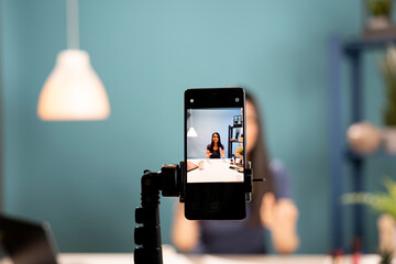 A smartphone camera captures a beauty influencer reviewing cosmetics. Mobile device screen showing asian vlogger using expressive gestures as she explains beauty products in a blue background studio.