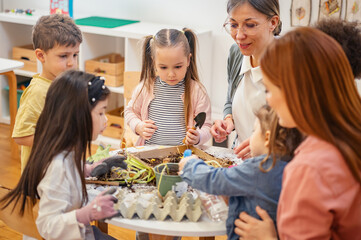 Group of diverse preschool children learning gardening with teachers, exploring soil and plants in...