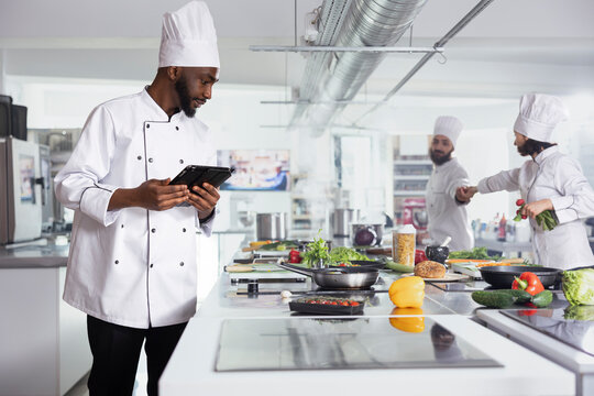 Black head chef in a restaurant kitchen uses laptop to refine recipes, while kitchen tools and fresh ingredients fill the workspace for gourmet food prep. Culinary arts for researching dishes.