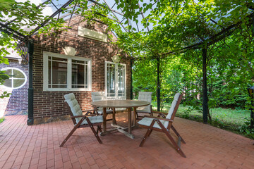 A patio with a shady, ivy-covered gazebo with garden furniture, on a sunny summer day.