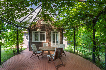 A cozy courtyard in the backyard of a brick house. A shady, ivy-covered gazebo with garden...