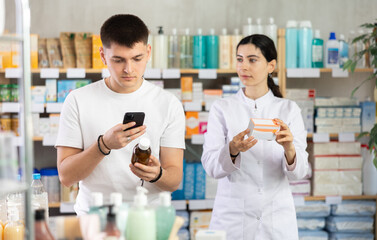 Obraz premium Near display case in pharmacy, guy scans barcode on antitussive syrup packaging, reads user manual. Client studies product in detail, collects information online. Girl pharmacist offer help
