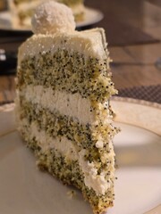 A tempting slice of cake features alternating layers of light green, poppy seed-speckled sponge and creamy white frosting, topped with a decorative white truffle.
