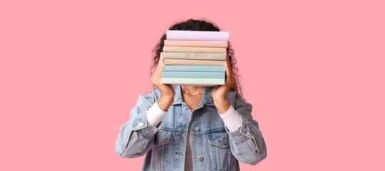 Young African-American woman hiding face behind stack of books on pink background