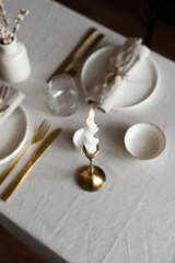 Spring minimalist table setting in warm neutral colors. Easter festive table decoration, white plates, gold cutlery, candles, eggs and willow branches in vase
