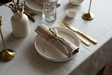 Spring minimalist table setting in warm neutral colors. Easter festive table decoration, white plates, gold cutlery, candles, eggs and willow branches in vase