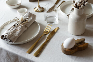 Spring minimalist table setting in warm neutral colors. Easter festive table decoration, white plates, gold cutlery, candles, eggs and willow branches in vase