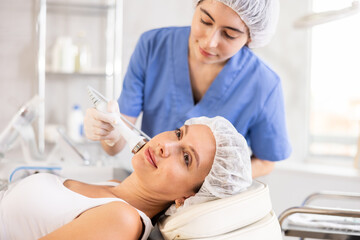 Obraz premium Young woman undergoing device-driven cosmetic facial treatment using high frequency ultrasound attachment stimulating opening of skin pores for deep penetration of moisturizers and nutrients