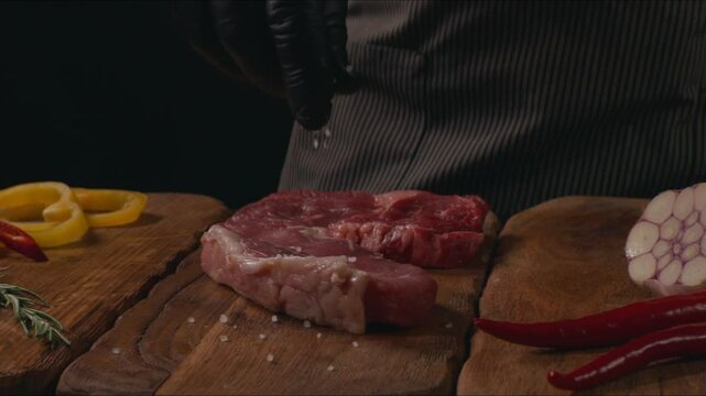 Chef preparing to cooking steak. Raw organic beef meat with rosemary, seasonings, salt and red pepper  on wooden cutting board, close-up. Slow motion