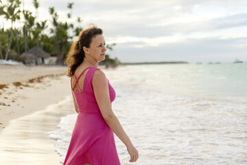 Woman in a pink dress strolls along a serene beach at sunrise near palm trees and calm ocean waves