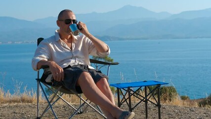 Tourist relaxing and drinking coffee by the sea. Man is enjoying a moment of relaxation by the sea, comfortably seated in a camping chair, sipping coffee from a metal cup and taking in the scenic view