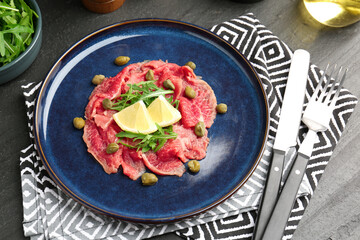 Fresh beef carpaccio with arugula, lemon and capers on dark textured table, above view