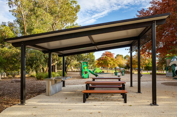 Modern outdoor picnic shelter with sleek black pergola roof, built-in barbecue facilities, and public picnic tables surrounded by autumn foliage. Contemporary park pavilion in a suburb of Australia.