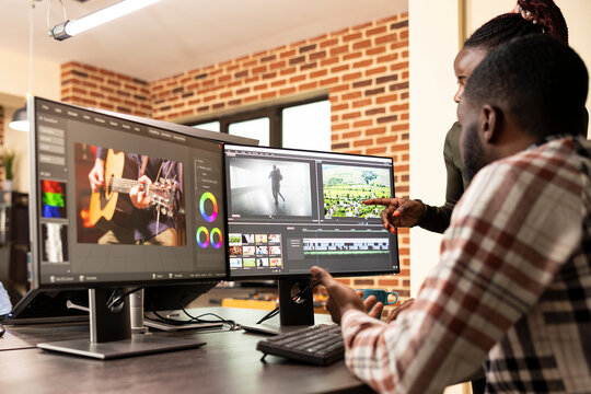 Professional male editor refining frames as coworker provides visual improvement support. Black man improving film quality at multi monitor workstation with assistance from female creative manager.