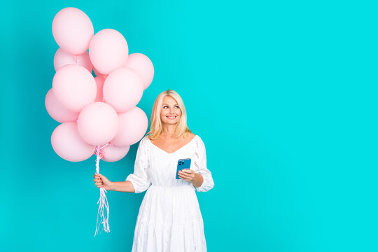 Smiling blonde woman holding pink balloons and smartphone wearing white dress on teal background, ready for celebration or photo shoot